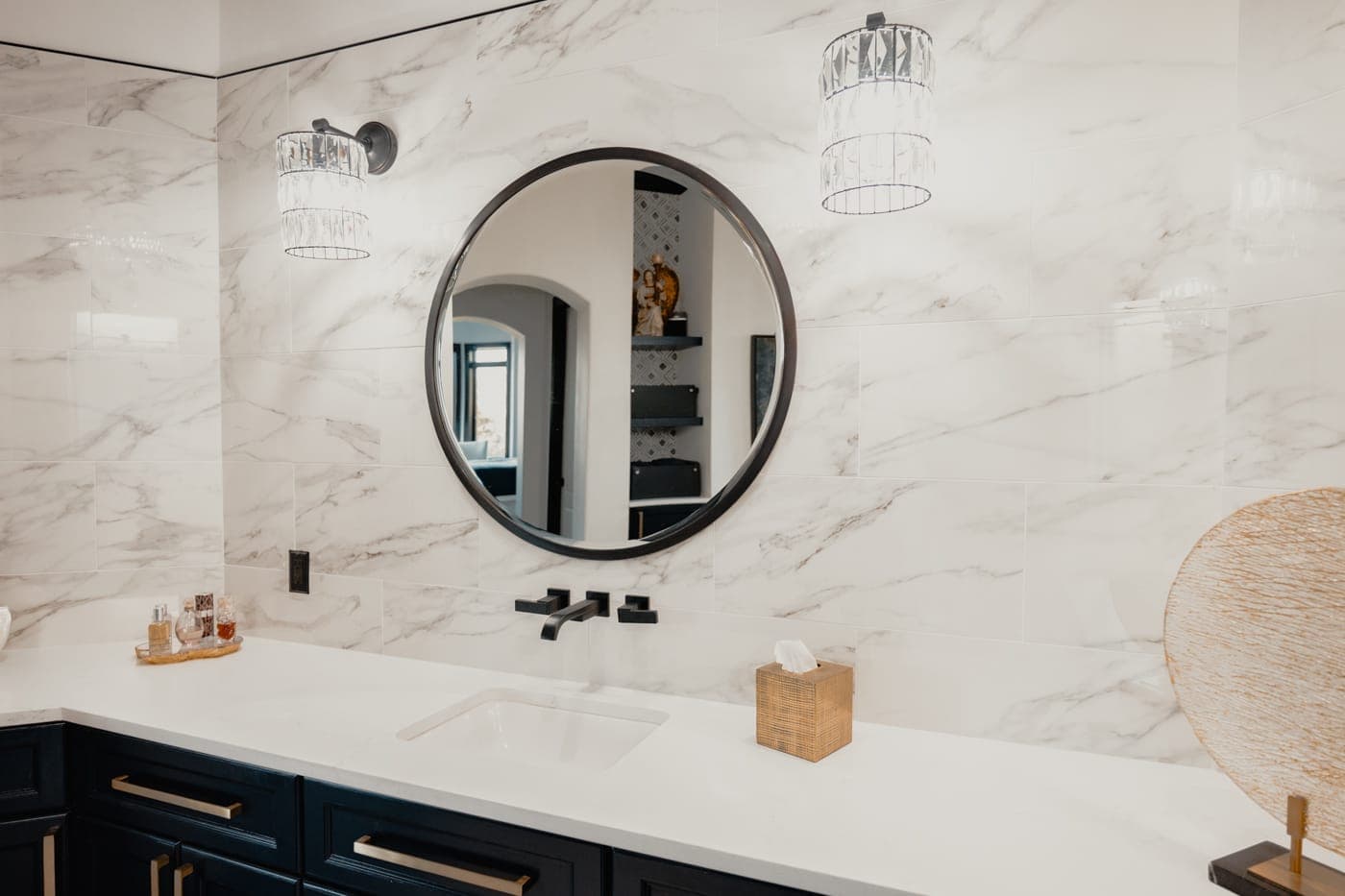 Round black-framed mirror over a quartz vanity against marble-look wall tile