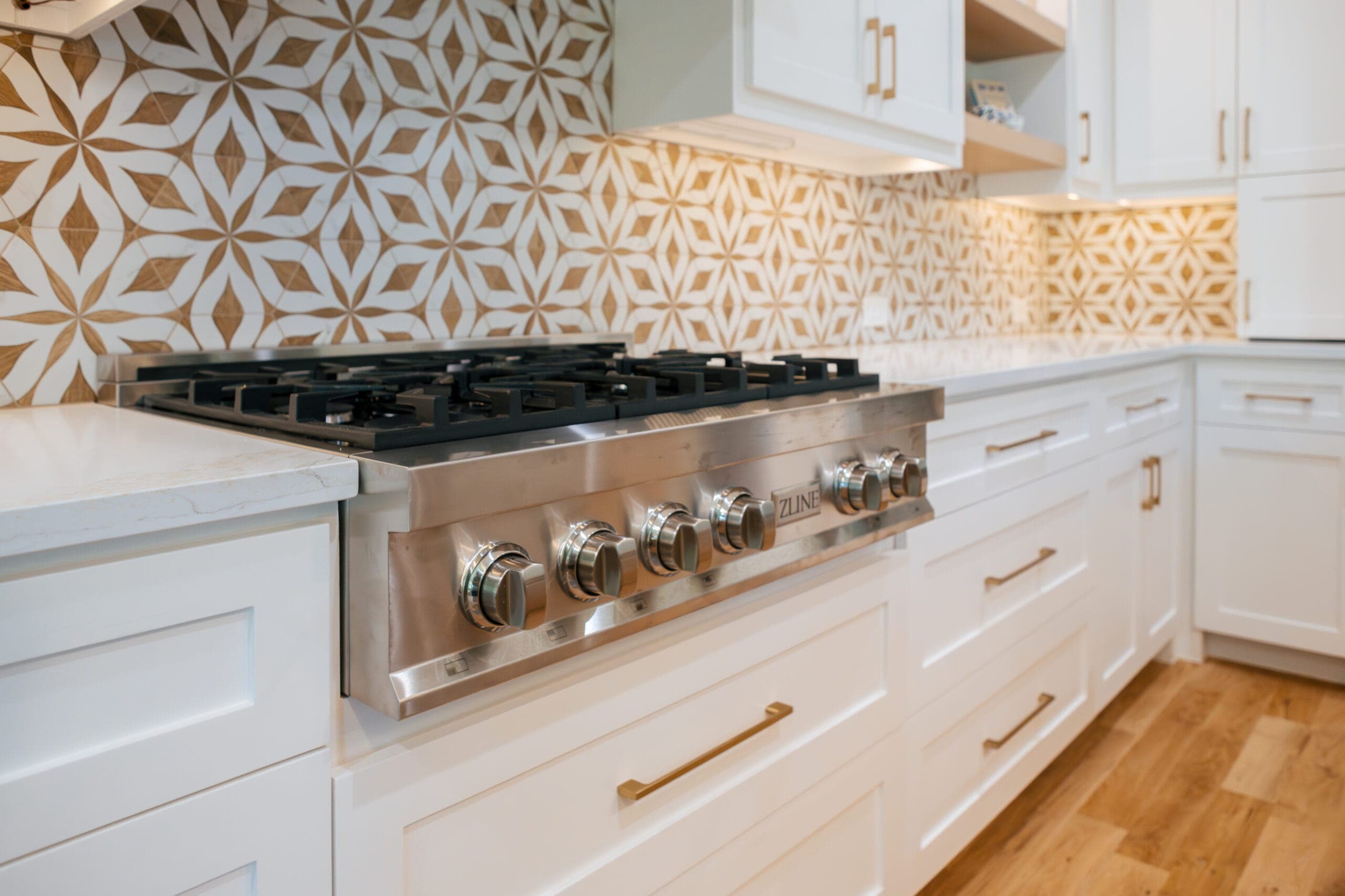 Stainless gas rangetop in a run of white shaker cabinets beneath a patterned tile backsplash