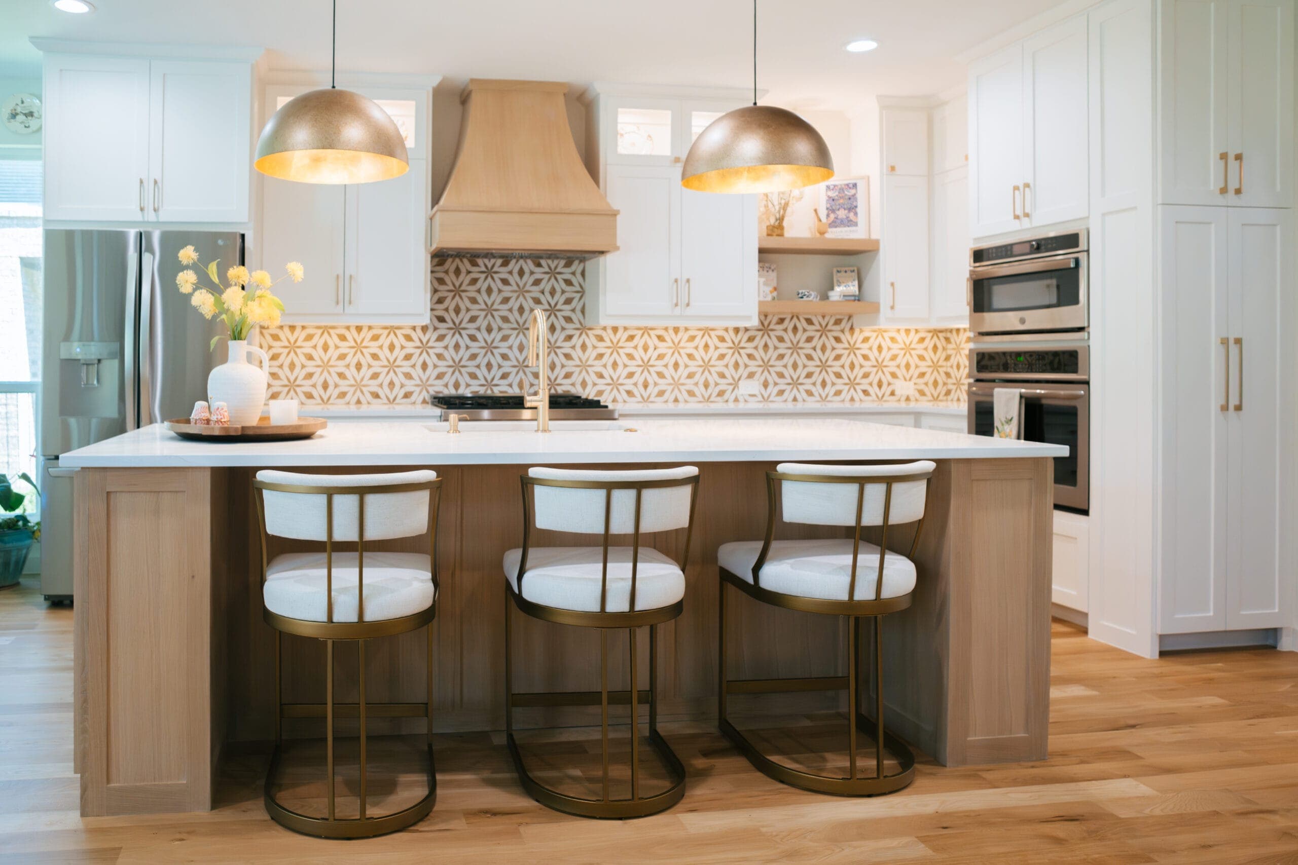 Three upholstered counter stools at a white oak island under two brass dome pendants — Southlake Luxury Kitchens, serving Southlake, TX